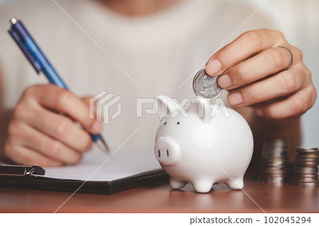Hand saving a coin into piggy bank, planning and writing information on paper with blurred coins and money on the wooden 102045294