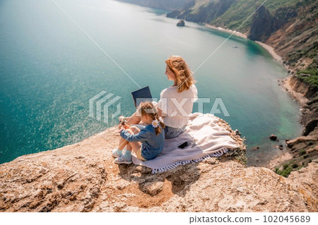Freelance woman with her daughter working on a laptop by the sea, typing on the keyboard, enjoying the beautiful view, highlighting the idea of remote work. Freelance woman with her daughter working on a laptop by the sea, typing on the keyboard, enjoying the beautiful view, highlighting the idea of remote work. 102045689