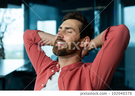 Close-up photo. Young man freelancer, student sits on a chair in the office, coffee shop and rests. He put his hands behind his head, closed his eyes. relaxes, meditates, dreams. 102048378