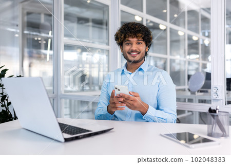 A young Latin American businessman sits in the office at a desk and uses a mobile phone, writes a message, searches for information on the Internet, checks social networks. Smiling at the camera. 102048383