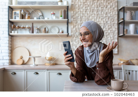 Close-up photo. Smiling Muslim young woman in hijab standing in kitchen at home, leaning on table and talking on mobile phone video call. 102048463