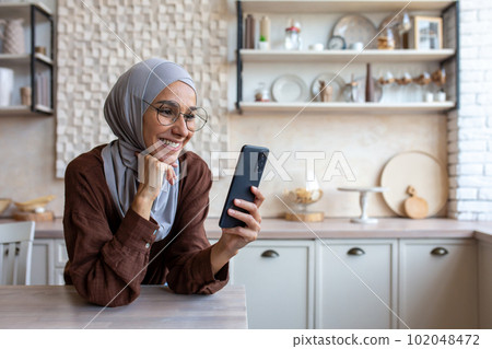 Close up photo. Smiling Muslim young woman in hijab standing in kitchen at home and using mobile phone, reading news, checking social media, watching videos, photos, searching for information. Close up photo. Smiling Muslim young woman in hijab standing in kitchen at home and using mobile phone, reading news, checking social media, watching videos, photos, searching for information. 102048472