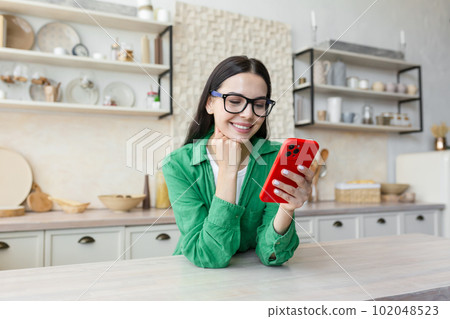 Young woman standing in kitchen at home, leaning on table and using mobile phone, chatting, checking social networks, sending messages, texting with friends, boyfriend. Young woman standing in kitchen at home, leaning on table and using mobile phone, chatting, checking social networks, sending messages, texting with friends, boyfriend. 102048523