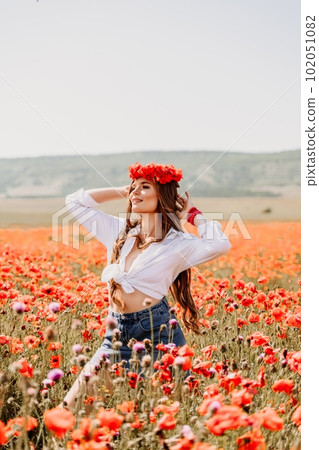 Happy woman in a poppy field in a white shirt and denim skirt with a wreath of poppies on her head posing and enjoying the poppy field. 102051082
