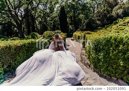 Brunette runs white dress park. A beautiful woman with long brown hair and a long white dress runs along the path along the beautiful bushes in the park, rear view Brunette runs white dress park. A beautiful woman with long brown hair and a long white dress runs along the path along the beautiful bushes in the park, rear view 102051091