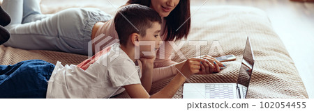 Small Caucasian cute boy with beautiful mother lying together on bed with laptop. 102054455