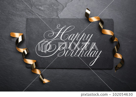 Slate board with greeting HAPPY BIRTHDAY and serpentine streamers on black background, top view 102054630