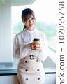 A young female worker drinking coffee and refreshing by the window in the office overlooking greenery 102055258