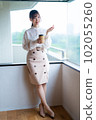 A young female worker drinking coffee and refreshing by the window in the office overlooking greenery 102055260