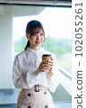 A young female worker drinking coffee and refreshing by the window in the office overlooking greenery 102055261