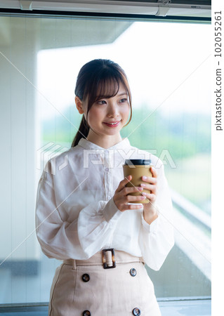 A young female worker drinking coffee and refreshing by the window in the office overlooking greenery A young female worker drinking coffee and refreshing by the window in the office overlooking greenery 102055261