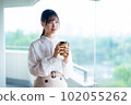 A young female worker drinking coffee and refreshing by the window in the office overlooking greenery 102055262