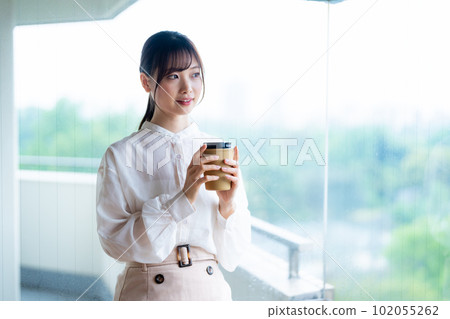 A young female worker drinking coffee and refreshing by the window in the office overlooking greenery A young female worker drinking coffee and refreshing by the window in the office overlooking greenery 102055262