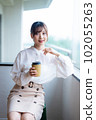 A young female worker drinking coffee and refreshing by the window in the office overlooking greenery 102055263