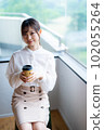 A young female worker drinking coffee and refreshing by the window in the office overlooking greenery 102055264