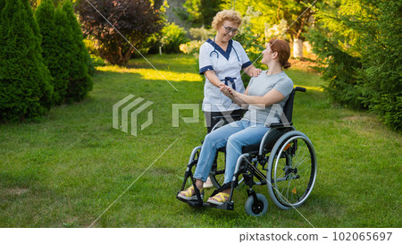 An elderly female nurse walks with a middle-aged woman in a wheelchair in the park. An elderly female nurse walks with a middle-aged woman in a wheelchair in the park. 102065697