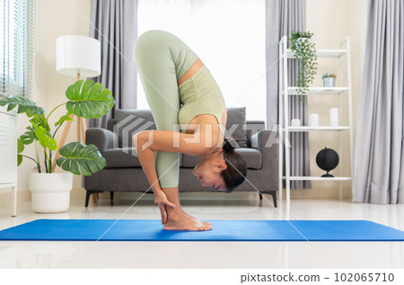 Side view of slim asian woman in green top and leggings doing yoga in standing forward fold or Uttanasana on blue mat in living room at home.. 102065710