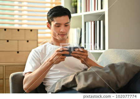 Happy millennial man playing video games on smart phone while resting on couch.Technology, entertainment and leisure activity Happy millennial man playing video games on smart phone while resting on couch.Technology, entertainment and leisure activity 102077774