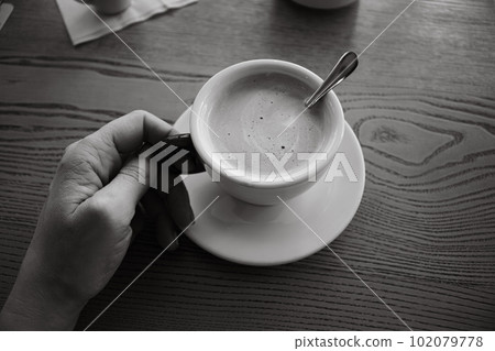 Woman hand holding coffee cup, black and white. Coffee with milk and sugar on table, monochrome. Morning drinks. Girl with cappuccino cup on table, top view. Coffee break concept. Woman hand holding coffee cup, black and white. Coffee with milk and sugar on table, monochrome. Morning drinks. Girl with cappuccino cup on table, top view. Coffee break concept. 102079778
