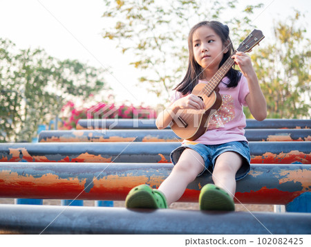 Little asian child girl play the ukulele, in the garden on the Steel pipe 102082425
