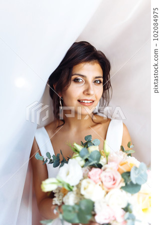 Young bride in white dress with a bouquet of flowers posing under curtain. Morning of the bride. 102084975