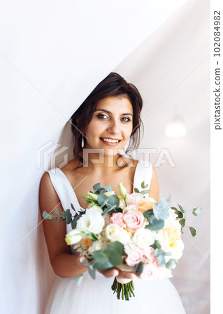 Young bride in white dress with a bouquet of flowers posing under curtain. Morning of the bride. Young bride in white dress with a bouquet of flowers posing under curtain. Morning of the bride. 102084982