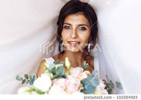 Young bride in white dress with a bouquet of flowers posing under curtain. Morning of the bride. Young bride in white dress with a bouquet of flowers posing under curtain. Morning of the bride. 102084985