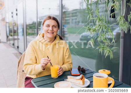 Happy woman sitting on the cafe terrace on the modern city street. 30s Person sitting at table and drinking drinks freshly squeezed orange fresh juice through straw in glass. Concept of easy breakfast 102085669