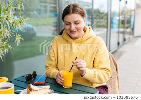 Happy woman sitting on the cafe terrace on the modern city street. 30s Person sitting at table and drinking drinks freshly squeezed orange fresh juice through straw in glass. Concept of easy breakfast Happy woman sitting on the cafe terrace on the modern city street. 30s Person sitting at table and drinking drinks freshly squeezed orange fresh juice through straw in glass. Concept of easy breakfast 102085670