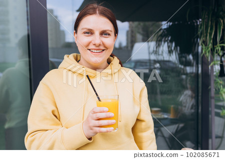 Happy woman sitting on the cafe terrace on the modern city street. 30s Person sitting at table and drinking drinks freshly squeezed orange fresh juice through straw in glass. Concept of easy breakfast Happy woman sitting on the cafe terrace on the modern city street. 30s Person sitting at table and drinking drinks freshly squeezed orange fresh juice through straw in glass. Concept of easy breakfast 102085671