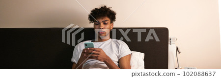 Young guy with piercing chatting with friends, using smartphone in his bed at night before going to sleep 102088637