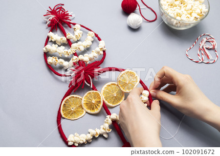 Female hands make Survakane, Survaknitsa, Survachka, Surva. Bulgarian Traditional Christmas Cornel Tree Twig. Flat lay with Popcorn, Red White Threads, Dried Orange. World Tree. Horizontal plane 102091672