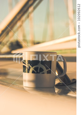 White cup with drawn helm on deck of a white yacht, ship, boat. Concept of tea coffee drinking while traveling at sea ocean. A mug in morning sunlight. Nautical wallpaper. Travel around the world. 102092612
