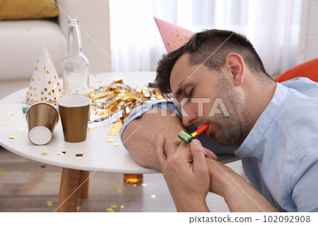 Young man in festive cap sleeping at table in room after party Young man in festive cap sleeping at table in room after party 102092908