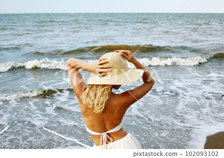 Blonde Woman in White Summer Style Standing at Sea and Holding Hat 102093102