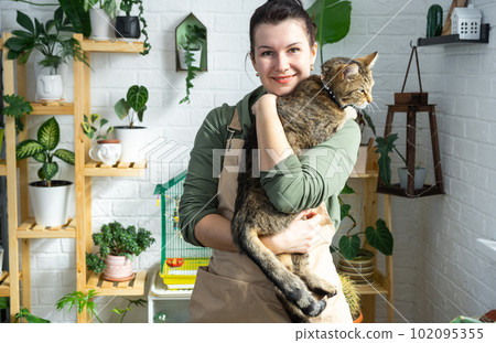 Woman holding pet cat in her arms near her collection of rare species home potted plants on shelf in interior of house. Home plant growing, green house, natural living corner Woman holding pet cat in her arms near her collection of rare species home potted plants on shelf in interior of house. Home plant growing, green house, natural living corner 102095355