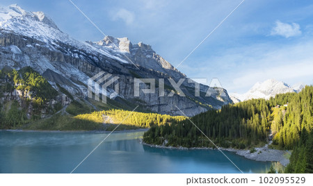 Idyllic morning view of the lake Oeschinensee. Location Swiss alps, Switzerland, Kandersteg district. Blue mountain lake with pine trees and mountains in background. Idyllic morning view of the lake Oeschinensee. Location Swiss alps, Switzerland, Kandersteg district. Blue mountain lake with pine trees and mountains in background. 102095529