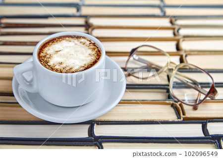 A white cup of fragrant cappuccino coffee and reading glasses on the background of worn old books folded upside down 102096549