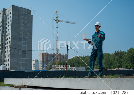 A male builder in a helmet stands against the background of a construction site with a laptop in his hands.  102112851
