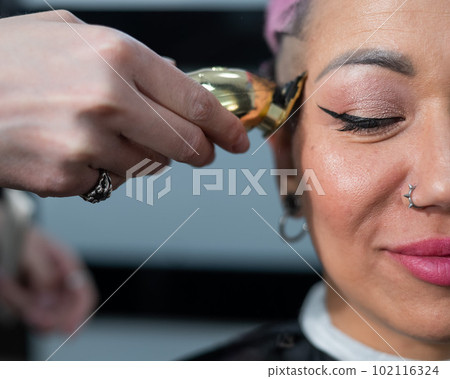 The hairdresser shaves the temple of a female client. Asian woman with short pink hair in barbershop. 102116324