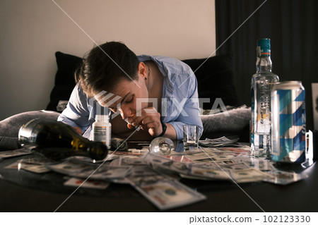 Young addicted junkie man sniffing snorting cocaine lines on mirror table with a lot of scattered money, alcohol, cards and pills through rolled banknote. Narcotics and addiction concept. 102123330