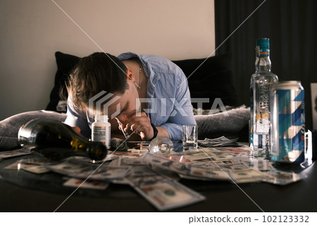 Young addicted junkie man sniffing snorting cocaine lines on mirror table with a lot of scattered money, alcohol, cards and pills through rolled banknote. Narcotics and addiction concept. 102123332