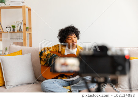 Blogger guitarist. African american girl blogger playing guitar talking to webcam recording vlog. Social media influencer woman streaming at home indoors. Music content creator broadcast tutorial 102124088