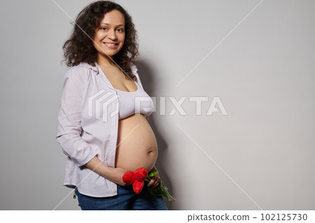 Happy pregnant woman, future mom holds bouquet of red tulips on her naked belly, smiles looking at camera, isolated white background. Happy Mothers Day. International Women's Day. Pregnancy 30 weeks 102125730
