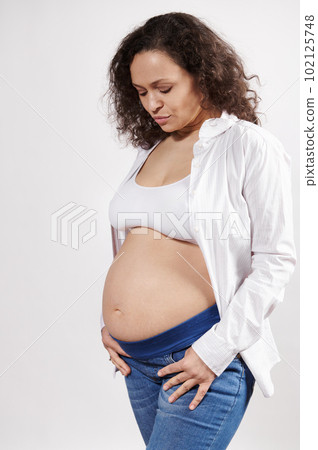 Vertical studio portrait of an attractive ethnic pregnant woman in jeans and white unbuttoned shirt, posing with a bare belly, isolated on a white background. Pregnant fashion. Body positivity 102125748