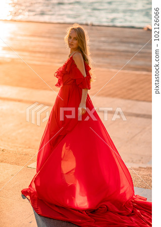 Sunrise red dress. A woman in a long red dress against the backdrop of sunrise, bright golden light of the sun's rays. The concept of femininity, harmony. 102126066
