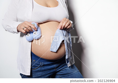 Close-up of gravid woman expecting baby, holding knitted booties and baby bodysuit over her bare belly, isolated on white background. Pregnant female with stretch marks on her stomach. Body positivity Close-up of gravid woman expecting baby, holding knitted booties and baby bodysuit over her bare belly, isolated on white background. Pregnant female with stretch marks on her stomach. Body positivity 102126904
