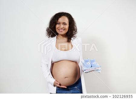 Attractive African American pregnant woman holding newborn clothes in her hand, gently caressing her belly, smiling looking at camera, isolated on white background with copy space. Pregnancy 30 weeks 102126935