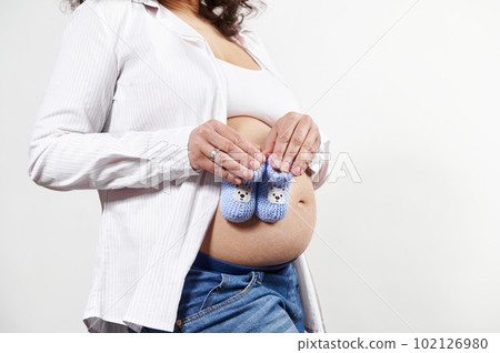 Close-up of a young pregnant woman, wearing jeans and unbuttoned white shirt, holding blue knitted baby booties on her naked belly, isolated on white background. Newborn clothing. Pregnancy. Maternity Close-up of a young pregnant woman, wearing jeans and unbuttoned white shirt, holding blue knitted baby booties on her naked belly, isolated on white background. Newborn clothing. Pregnancy. Maternity 102126980