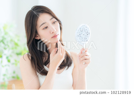 A young woman doing skin care while looking at a hand mirror in the living room A young woman doing skin care while looking at a hand mirror in the living room 102128919